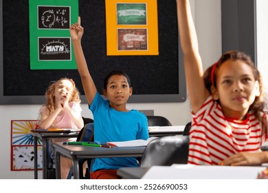 Diverse students raise hands in classroom. A biracial boy in a blue shirt, a biracial girl in a red-striped shirt, and a Caucasian girl with blonde hair participate, unaltered - Powered by Shutterstock