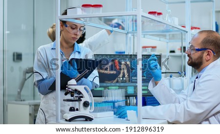 Diverse team of biochemistry scientists developing drugs agains new virus, doctor checking samples while nurse taking noters on clipboard in modern equipped laboratory. Multiethnic scientists working