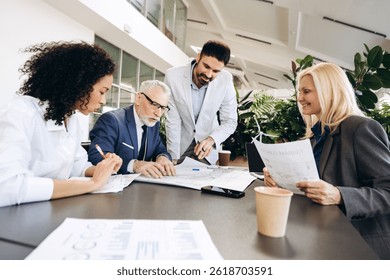 Diverse team of business professionals collaborating on sustainable development project, analyzing blueprints, market reports, and financial data in modern office environment. Meeting, startup concept