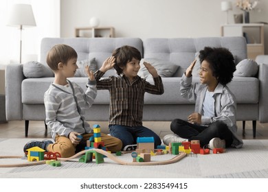 Diverse team of cheerful kids playing game on home floor, constructing towers, city road from toy building bricks, giving group high five, celebrating teamwork success - Powered by Shutterstock