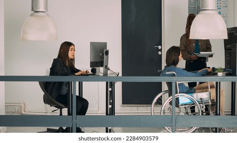 Diverse team of cloud programers working on app developing, it engineer in wheelchair talking to coder in company office. Database admin with impairment creating firewall security server.