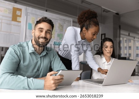 Diverse team collaborate on task gather in conference room, focus on smiling Hispanic man sitting at desk with digital tablet, look at camera exuding leadership and satisfaction, using innovative tech