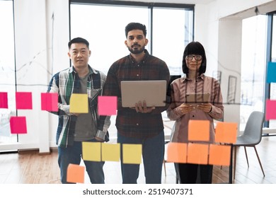 Diverse team collaborating in modern office environment using sticky notes on glass wall. Team members with laptops and tablets planning and brainstorming. Modern workspace with city view. - Powered by Shutterstock