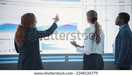 Diverse Team Of Data Scientists Analyzing Information And Graphs On Big Digital Screen In Modern Monitoring Office. Multiethnic Colleagues Discussing Financial Strategies, Risk Factors, and Deals.