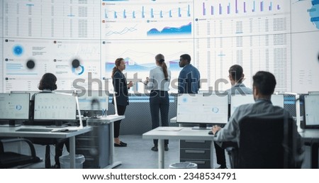 Diverse Team Of Data Scientists Analyzing Information And Graphs On Big Digital Screen In Modern Monitoring Office. Multiethnic Employees Working Infront Of Desktop Computers In Consulting Firm.