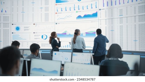 Diverse Team Of Data Scientists Having A Meeting And Discussing Reports In a Modern Monitoring Office With Analytics Feed On a Big Digital Screen. Big Data Company With Employees Behind Computers