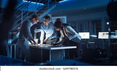 Diverse Team Of Electronics Development Engineers Standing At The Desk Working With Documents, Solving Project Problems Late At Night. Specialists Working On Ultra Modern Industrial Design.