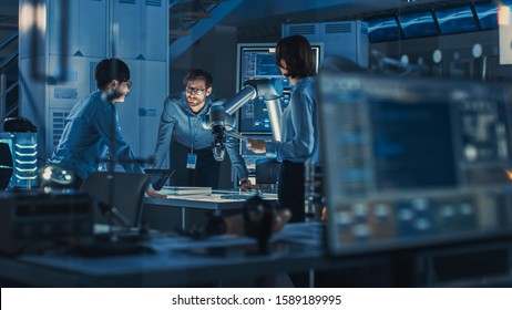 Diverse Team Of Engineers With Laptop And A Tablet Analyse And Discuss How A Futuristic Robotic Arm Works And Moves A Metal Object. They Are In A High Tech Research Laboratory With Modern Equipment.