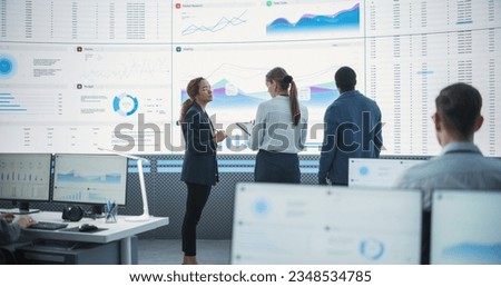 Diverse Team Of Financial Data Analysts Studying Information And Graphs On Big Digital Screen In Modern Monitoring Office. Multiethnic Employees Working Infront Of Desktop Computers In Consulting Firm