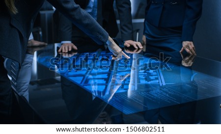 Diverse Team of Government Intelligence Agents Standing Around Digital Touch Screen Table and Satellite Tracking Suspect, Pointing at Display. Big Dark Surveillance Room.