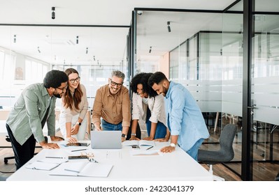 Diverse team huddles around table, and looks at a laptop screen in a bright office, closely reviewing documents and digital content together, showcasing a moment of collective focus and determination - Powered by Shutterstock