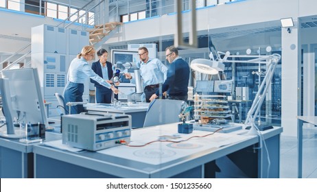 Diverse Team Of Industrial Robotics Design Engineers Gathered Around Table With Modern Moving Robot Arm. Specialists Talking, Programming And Manipulating Robot Hand. Bright High Tech Facility