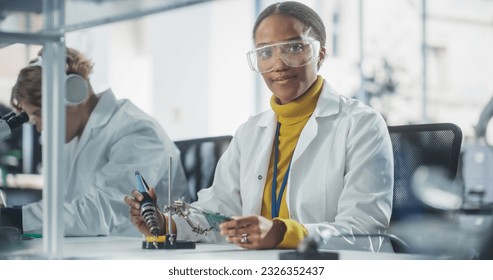 Diverse Team of Multiethnic Young Scientists Passing Internship in a Modern High Tech Laboratory. Cute African Female Looking at Camera and Smiling while Working with Soldering Iron - Powered by Shutterstock