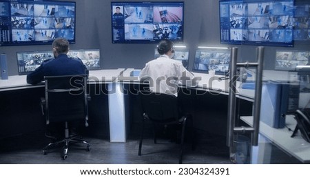 Diverse team of officers watch CCTV cameras with AI facial recognition. Employees work in security control center. Computer monitors and big digital screens showing surveillance cameras video footage.
