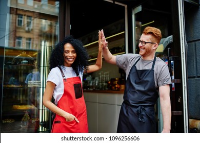 Diverse team of positive professional barista and experienced waiter in aprons giving five each other standing outdoors near own coffee shop.Cheerful young man and woman developing own cafe business