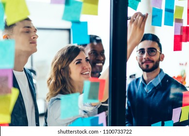 Diverse team of positive young people laughing while working together during brainstorming and standing behind glass wall with sticky colorful papers.Cheerful students learning words from stickers - Powered by Shutterstock