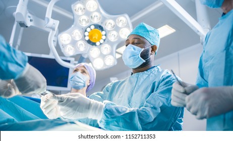 Diverse Team Of Professional Surgeon, Assistants And Nurses Performing Invasive Surgery On A Patient In The Hospital Operating Room. Surgeons Talk And Use Instruments. Real Modern Hospital.