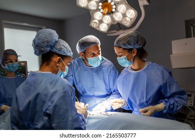 Diverse Team Of Professional Surgeon, Assistants And Nurses Performing Invasive Surgery On A Patient In The Hospital Operating Room. Surgeons Talk And Use Instruments. Real Modern Hospital.