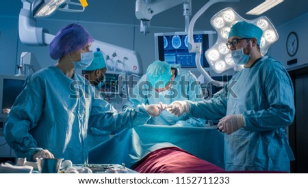 Diverse Team of Professional Surgeons Performing Invasive Surgery on a Patient in the Hospital Operating Room. Nurse Hands Out Instruments to surgeon, Anesthesiologist Monitors Vitals. Modern Hospital