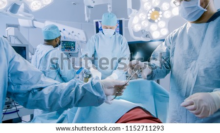 Diverse Team of Professional Surgeons Performing Invasive Surgery on a Patient in the Hospital Operating Room. Nurse Passes Instrument to a Chief Surgeon, Anesthesiologist Monitors Vitals.