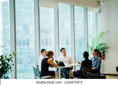 A Diverse Team Sit Around A Table In A Meeting Room To Have A Business Meeting To Discuss Plans. The Group Is International With Asian And White Team Members And They Are All Professionally Dressed.