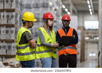 A diverse team of warehouse workers in safety gear reviews logistics with a tablet. The industrial background highlights the focus on teamwork, communication, and safety in a busy work environment. - Powered by Shutterstock