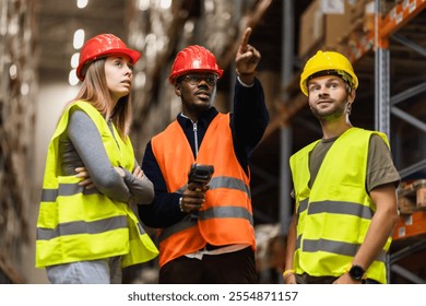 A diverse team of warehouse workers wearing safety vests and helmets discuss logistics. They appear focused, collaborating on tasks in a busy industrial environment. - Powered by Shutterstock