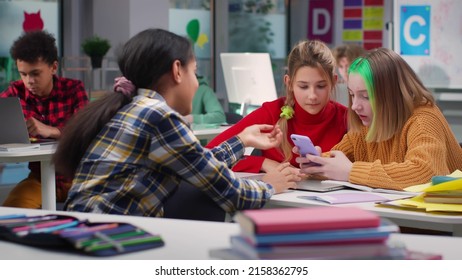 Diverse Teen Kids At School Discuss Content On Cellphone Sitting In Classroom. Girlfriends Chat And Gossip Looking At Smartphone Sitting In School Class