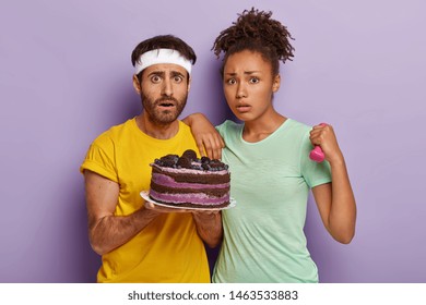 Diverse Unhappy Couple Do Sport Together, Look With Temptation At Delicious Cake, Afro Woman Holds Dumbbell, Want To Eat Something Sweet, Isolated On Purple Background. Sport And Unhealthy Eating