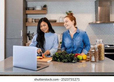 Diverse women in the kitchen with laptop watching online cooking classe. Live streaming cooking class, online class. Asian woman watching online class while cooking. Preparing food in kitchen. - Powered by Shutterstock