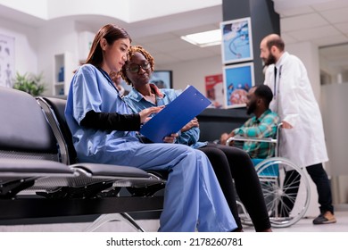 Diverse Women Talking About Healthcare Service At Clinic, Discussing About Treatment And Medicine In Waiting Room Area. Assistant Consulting Female Patient At Checkup Appointment.