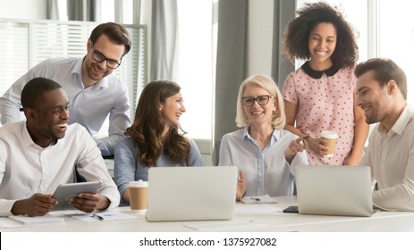 Diverse Workers Have Fun Watch Video Joke Online Use Pc Drink Coffee Break Sitting In Office Room Workplace, Different Ages Ethnicity Employees Members Laughing At Training With Mature Business Coach
