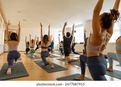 Diverse yoga class executes a kneeling arm stretch, enhancing balance and flexibility in a serene, sunlit studio with wooden flooring