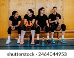 Diverse young beautiful girls sitting on a bench after training or playing volleyball match in a sports hall. Woman volleyball squad taking a break while resting and drinking water together.