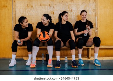 Diverse young beautiful girls sitting on a bench after training or playing volleyball match in a sports hall. Woman volleyball squad taking a break while resting and drinking water together. - Powered by Shutterstock