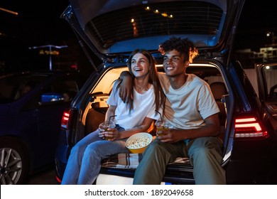 Diverse Young Couple Having Romantic Date. Cheerful Guy And His Girlfriend Watching A Movie, Sitting Together In Car Trunk In Front Of Screen In Open Air Cinema. Entertainment Ideas. Horizontal Shot