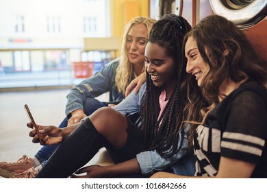 Diverse Young Female Friends Laughing While Sitting Together On The Floor Of A Laundromat Using A Cellphone