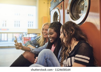 Diverse Young Female Friends Laughing And Sitting Together On A Laundromat Floor Using A Cellphone