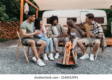 Diverse young friends sitting near campfire, telling scary stories next to motorhome during camping trip, spending summer vacation ourdoors. Travel and recreation in summertime