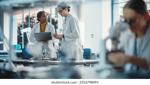 Diverse Young Group of Engineers Working in a Startup Lab. Female Specialist Checking on Electronic Circuit Using Microscope While Two Project Managers Discuss Manufacturing Using Laptop Computer - Powered by Shutterstock