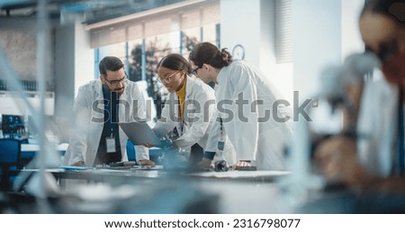 Diverse Young Group of Engineers Working in a Startup Company Lab. Project Managers Discuss Manufacturing Using Laptop Computer While Female Specialist Checking on Electronic Circuit Using Microscope