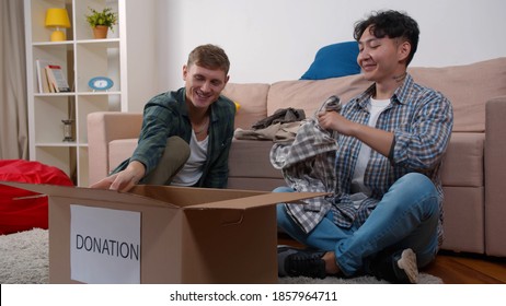 Diverse Young Men Volunteers Packing Cardboard Boxes With Clothing For Donation Or Recycling. Friends Or Gay Couple Putting Old Clothes In Donation Boxes For Social Support