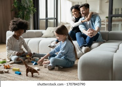 Diverse young parents sit on couch in living room watch multiracial daughters playing together, happy multiethnic family with little girls children have fun enjoy leisure quarantine weekend at home