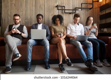 Diverse Young People Sitting In Row On Couch Together Obsessed With Devices Online, African And Caucasian Millennial Addicts Using Laptops And Smartphones, Digital Life And Gadgets Overuse Concept