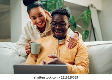 Diverse Young Relaxed Couple Using Laptop While Hugging And Drinking Coffee. Shopping Online Or Web Surfing. Multiracial Couple Spending Time At Home. Copy Space.