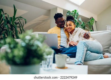 Diverse Young Relaxed Couple Using Laptop While Watching Funny Movie Or Enjoying Comedian Film And Hugging At Home On The Sofa. Shopping Online Or Web Surfing. Multiracial Couple Spending Time At Home