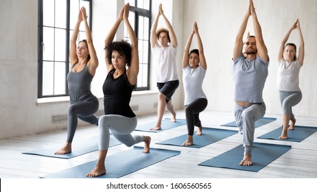 Diverse young sporty people doing Warrior one exercise at group lesson, practicing yoga in modern fitness center, standing in Virabhadrasana pose, working out with African American female instructor