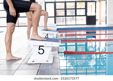 Diverse young swimmer competitors, including a Caucasian male swimmer standing on starting block at poolside indoors. He has short brown hair, wearing black swimwear, preparing for dive, unaltered - Powered by Shutterstock