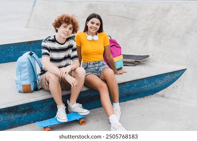 Diversity couple of hispanic girl caucasian redhead curly boy teenagers friends pupils classmates students in casual outfit with bags riding on skateboard sitting on skate ramp park looking at camera - Powered by Shutterstock