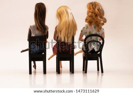 
Diversity. Three dolls of different colors and hair sitting on black chairs, seen from the back. White background.